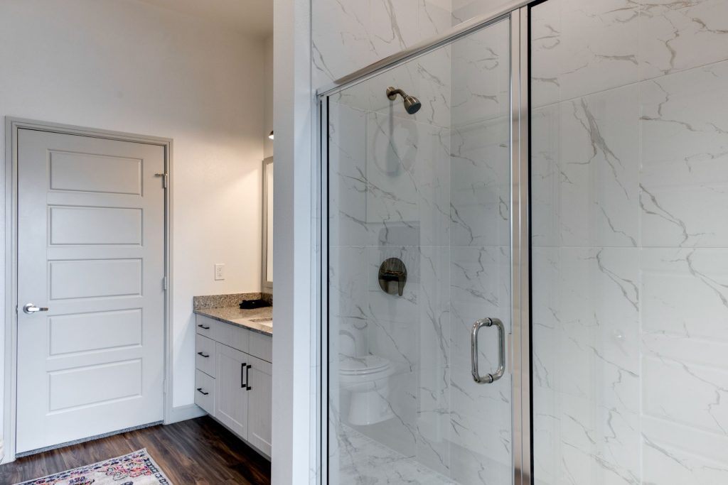 Bathroom area with large mirror, single sink, granite countertops, wood style flooring, standup shower, and cool lighting