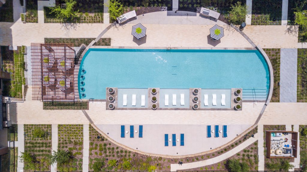 Aerial view of outdoor pool area with shaded seating, sun tan chairs, tropical landscaping, and grilling stations