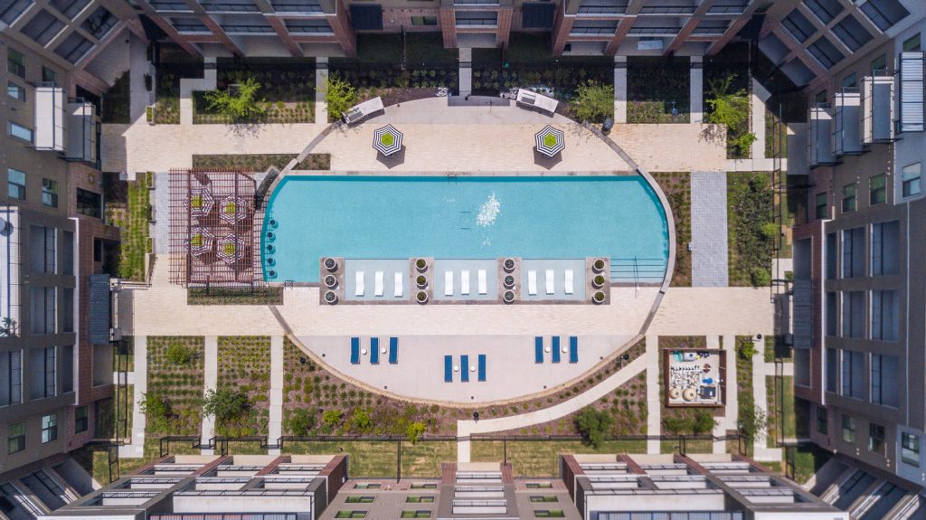 Aerial view of outdoor pool area with shaded seating, sun tan chairs, tropical landscaping, and grilling stations