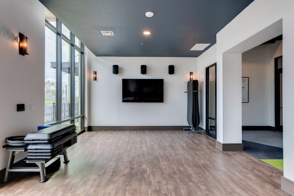 Empty indoor gym space for dance and yoga classes, with wall-to-wall mirror, yoga mats, television, and floor to ceiling windows