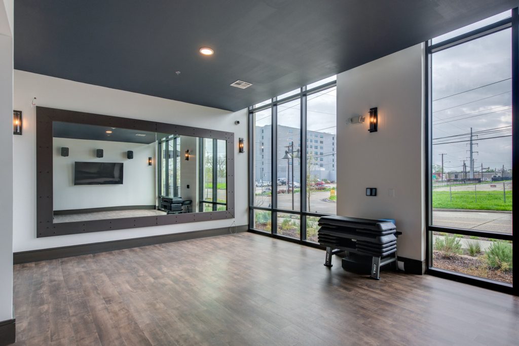 Empty indoor gym space for dance and yoga classes, with wall-to-wall mirror, yoga mats, television, and floor to ceiling windows