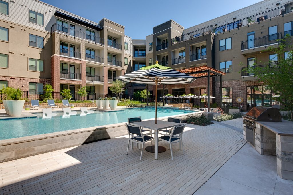 Outdoor pool area with shaded seating, sun tan chairs, tropical landscaping, and grilling stations