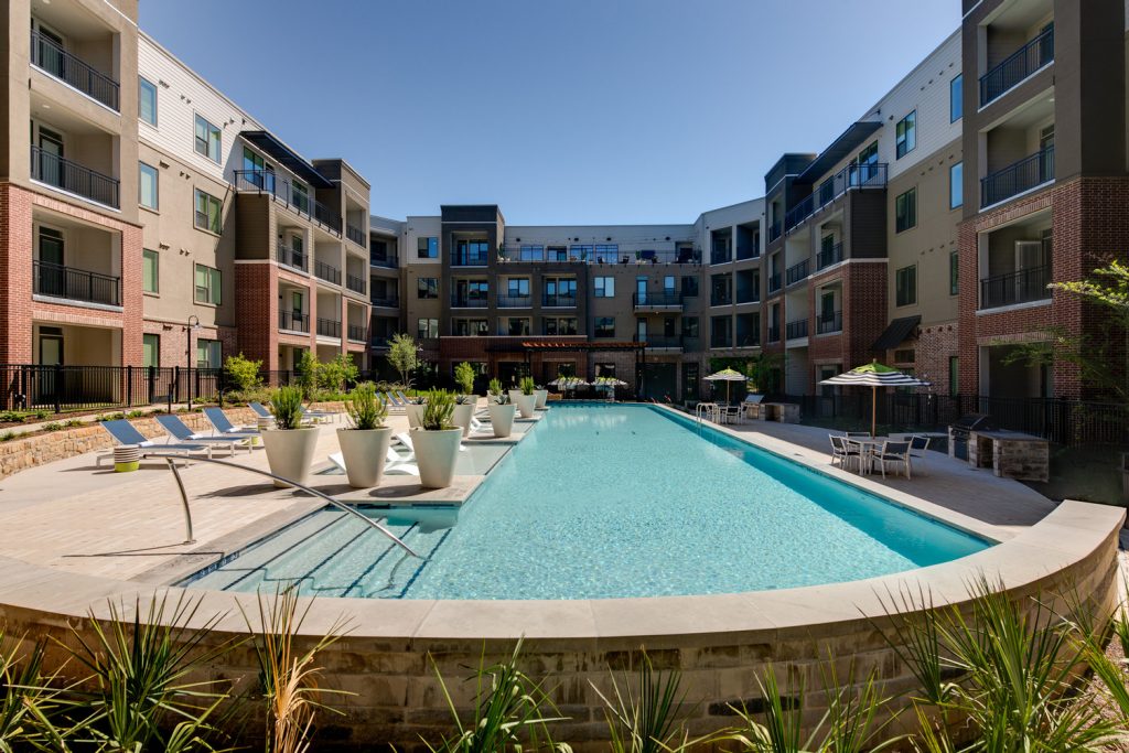 Outdoor pool area with shaded seating, sun tan chairs, tropical landscaping, and grilling stations