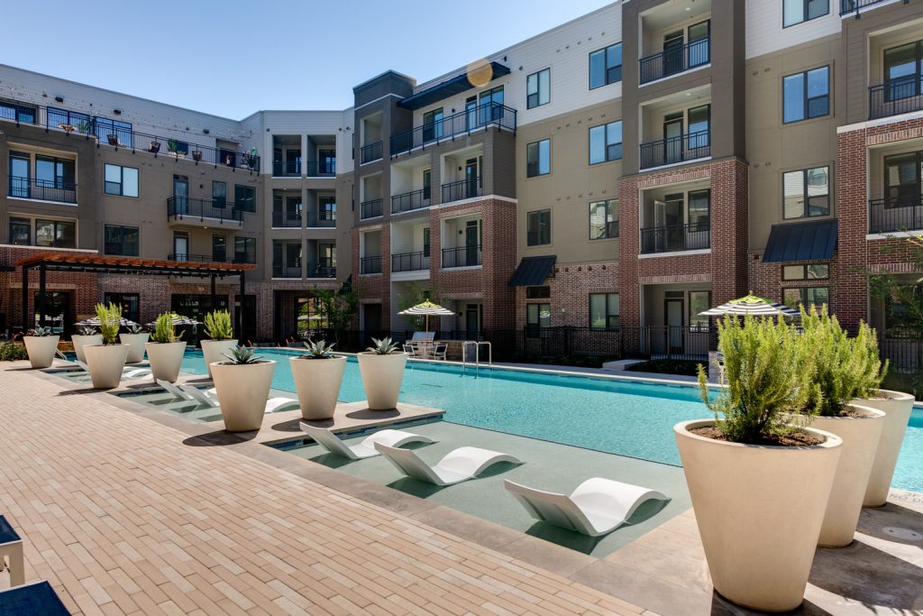 Outdoor pool area with shaded seating, sun tan chairs, tropical landscaping, and grilling stations