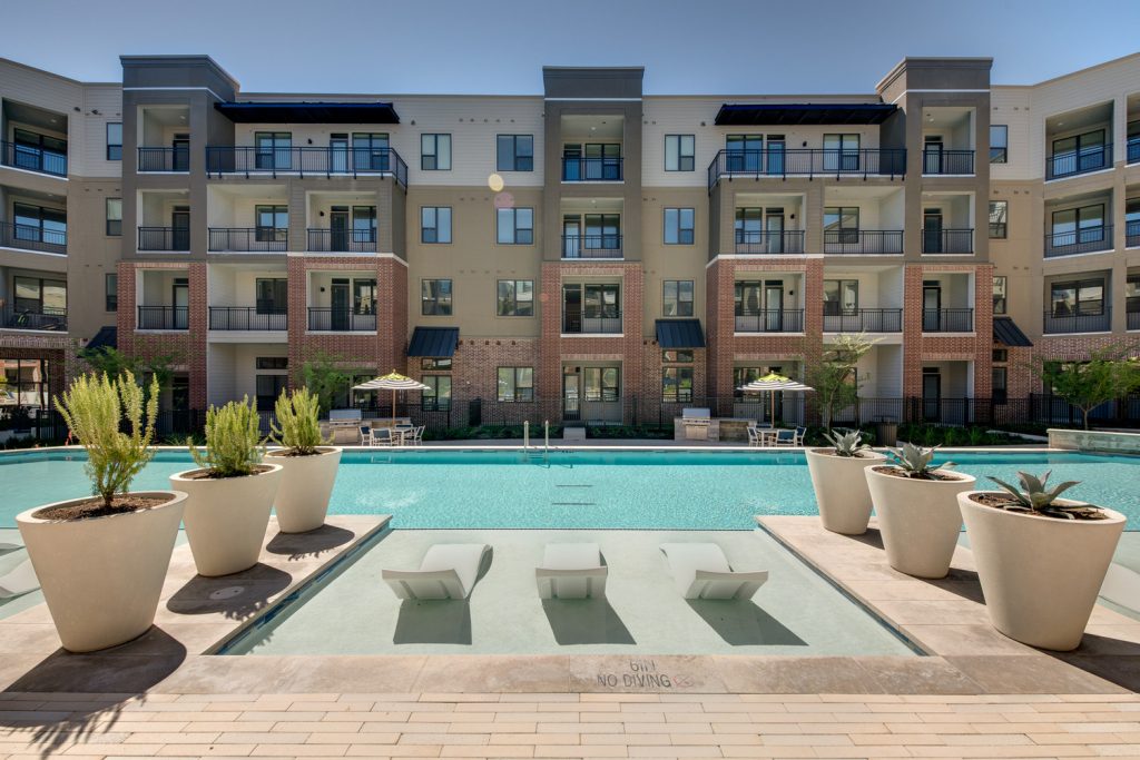 Outdoor pool area with shaded seating, sun tan chairs, tropical landscaping, and grilling stations