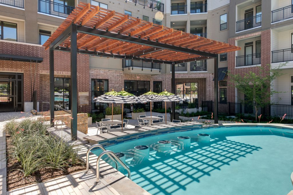 Outdoor pool area with shaded seating, sun tan chairs, tropical landscaping, and grilling stations