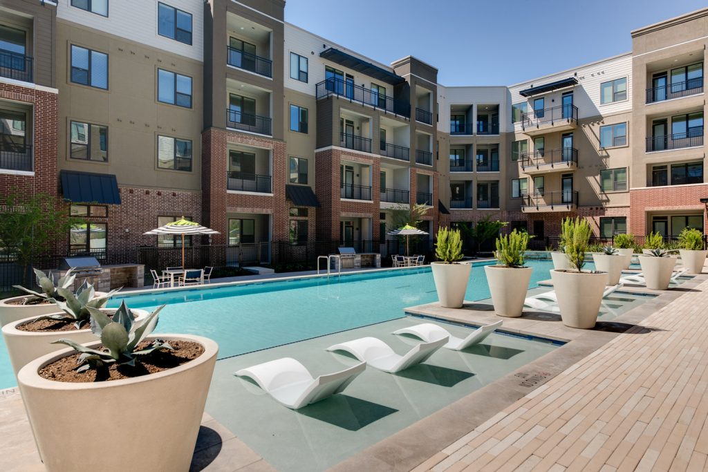 Outdoor pool area with shaded seating, sun tan chairs, tropical landscaping, and grilling stations