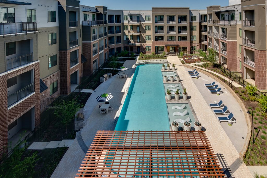 Aerial view of outdoor pool area with shaded seating, sun tan chairs, tropical landscaping, and grilling stations