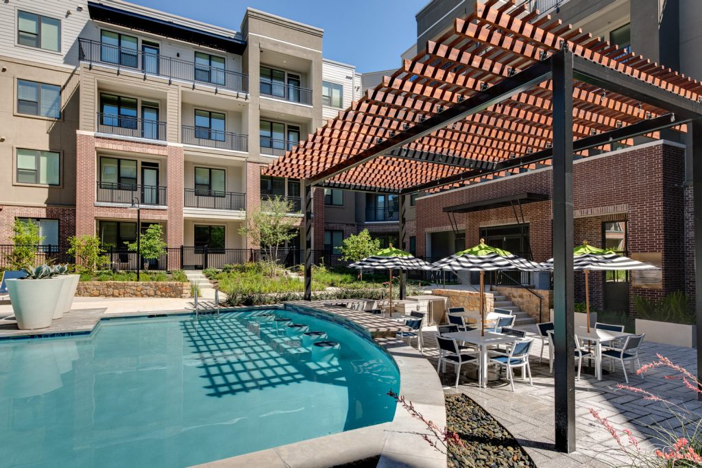 Outdoor pool area with shaded seating, sun tan chairs, tropical landscaping, and grilling stations