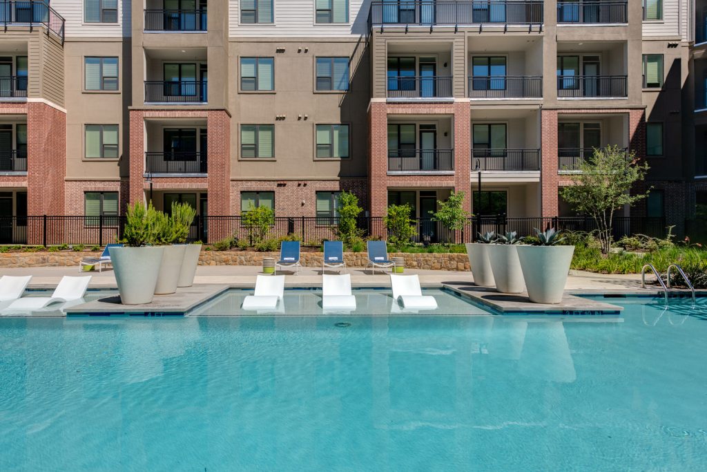 Outdoor pool area with shaded seating, sun tan chairs, tropical landscaping, and grilling stations