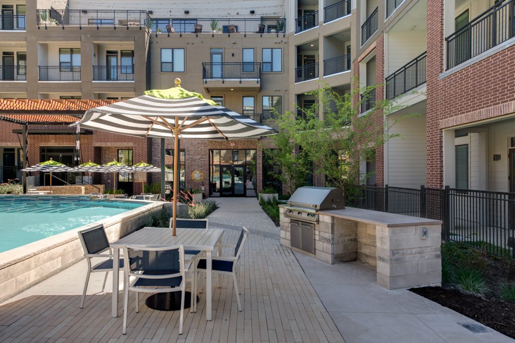 Outdoor pool area with shaded seating, sun tan chairs, tropical landscaping, and grilling stations