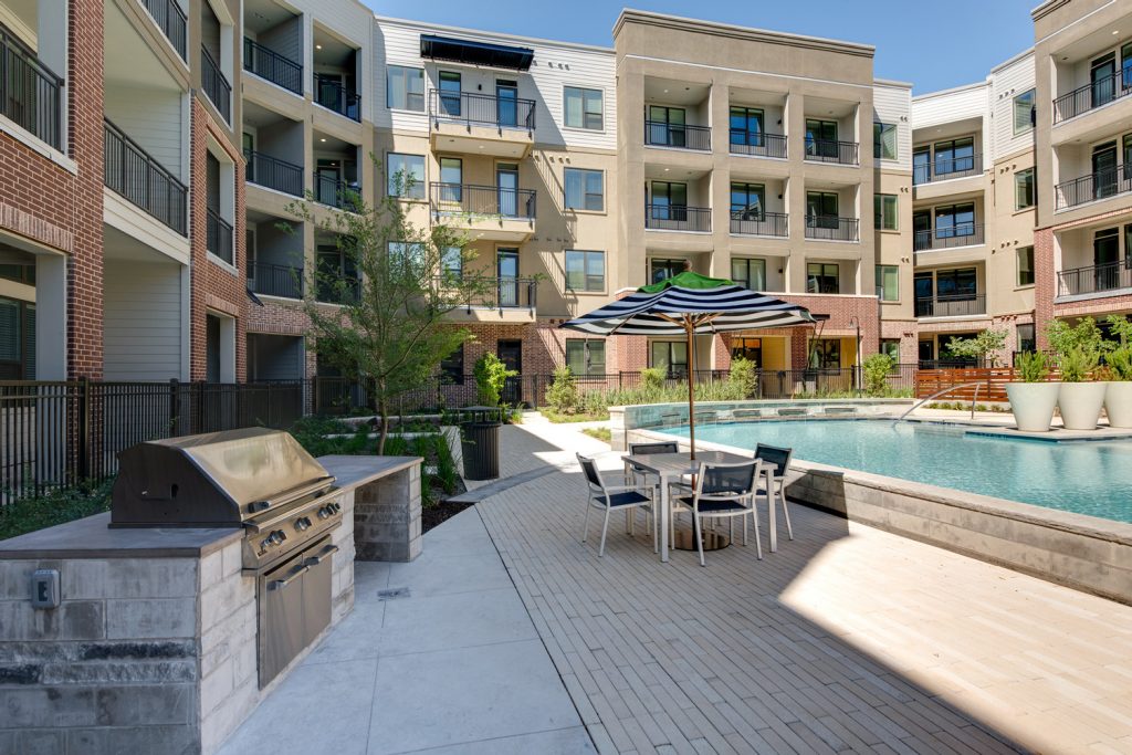 Outdoor pool area with shaded seating, sun tan chairs, tropical landscaping, and grilling stations